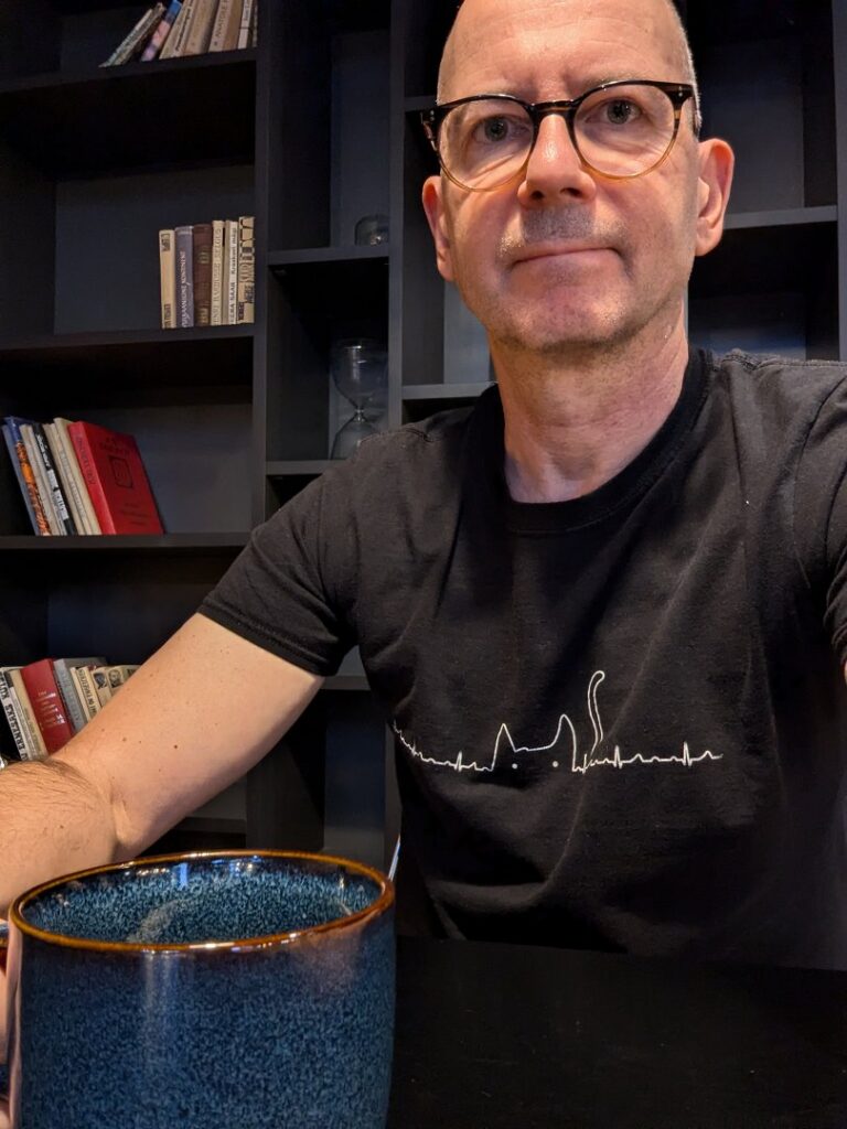 Mann in Tallinn mit Brille und Katze-T-Shirt. Bücherregal im Hintergrund. Kaffeetasse. Ibis-Hotel, Tallinn