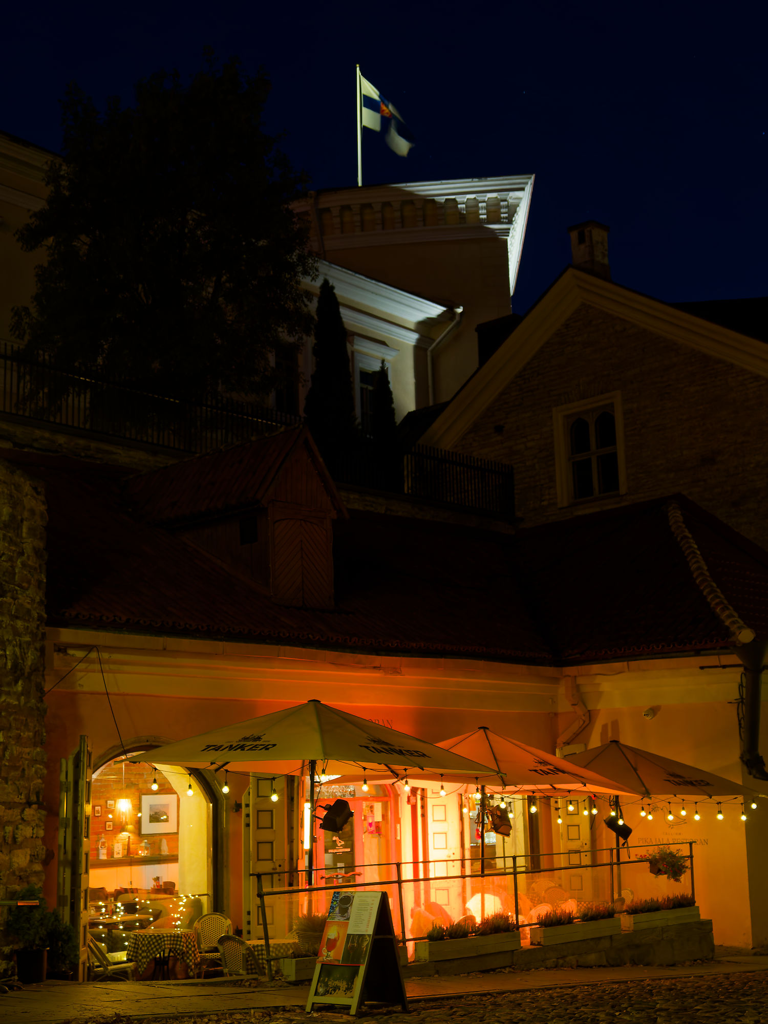 Nächtliches Tallinn: Gemütliches Café mit estnischer Flagge im Hintergrund. Stimmungsvolle Beleuchtung. Tallinn Altstadt.