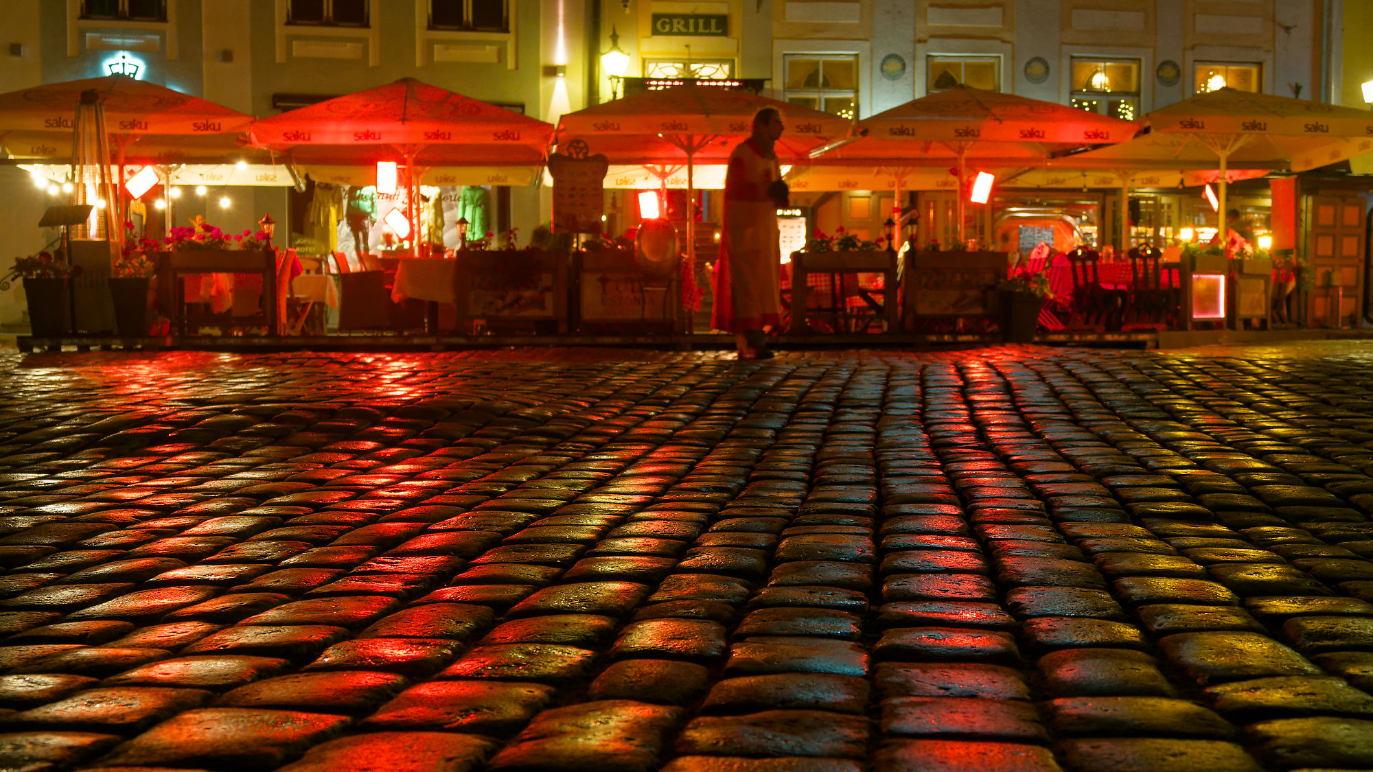 Tallinn bei Nacht: Pflastersteine und beleuchtete Restaurants mit Sonnenschirmen. Stimmungsvolle Stadtansicht.