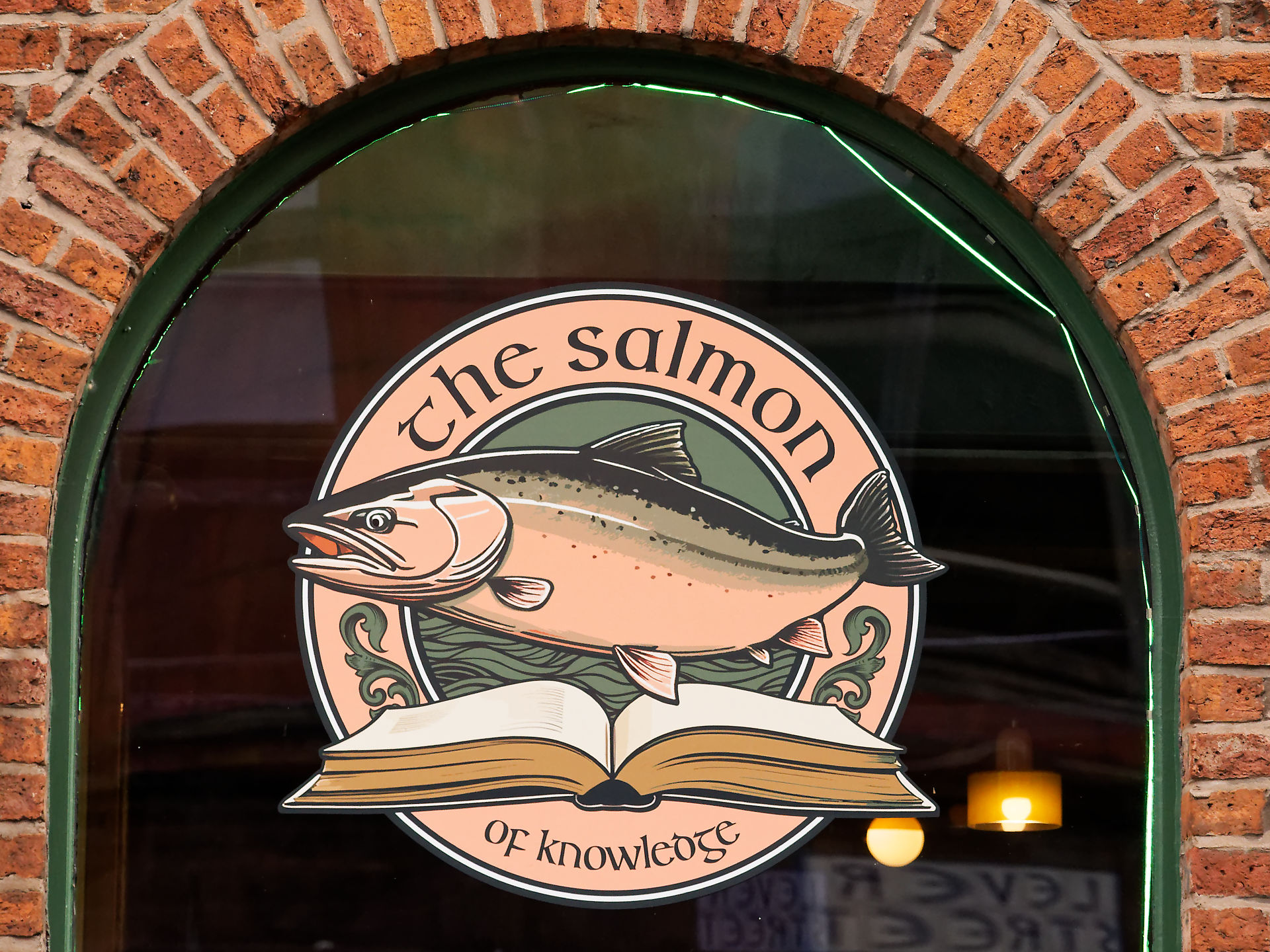 Manchester Das Lachs-Logo auf einem Buch, das "The Salmon of Knowledge" in einem Backsteinbogenfenster darstellt.