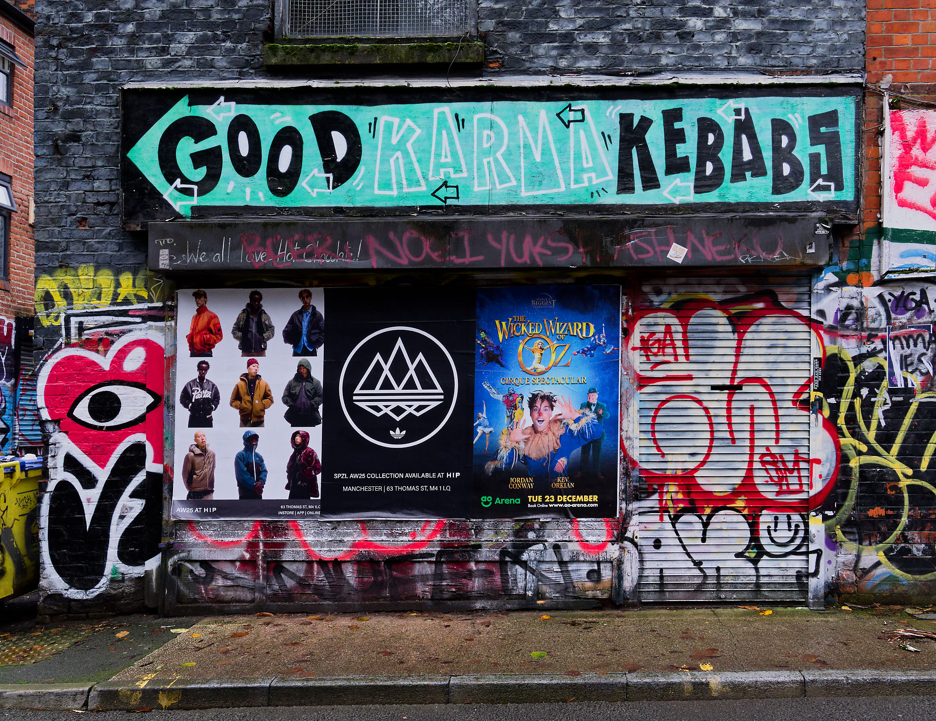 Manchester Manchester Street Scene: "Good Karma Kebabs" sign, graffiti art, Adidas poster, "Wicked Wizard of Oz" advertisement.