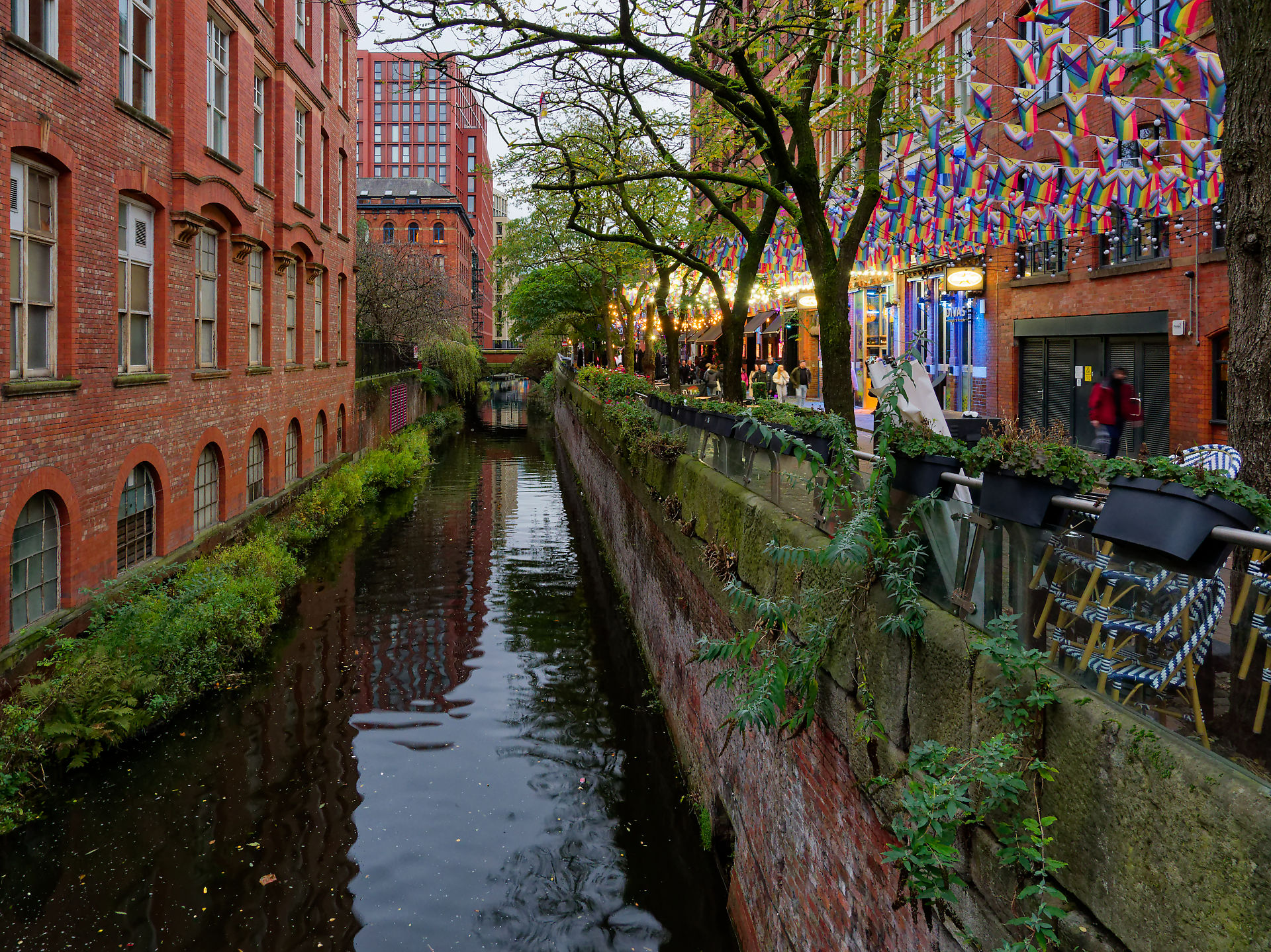 Manchester Manchester Kanalansicht mit roten Backsteingebäuden, Bäumen und bunten Flaggen. Stimmungsvolles Stadtbild.