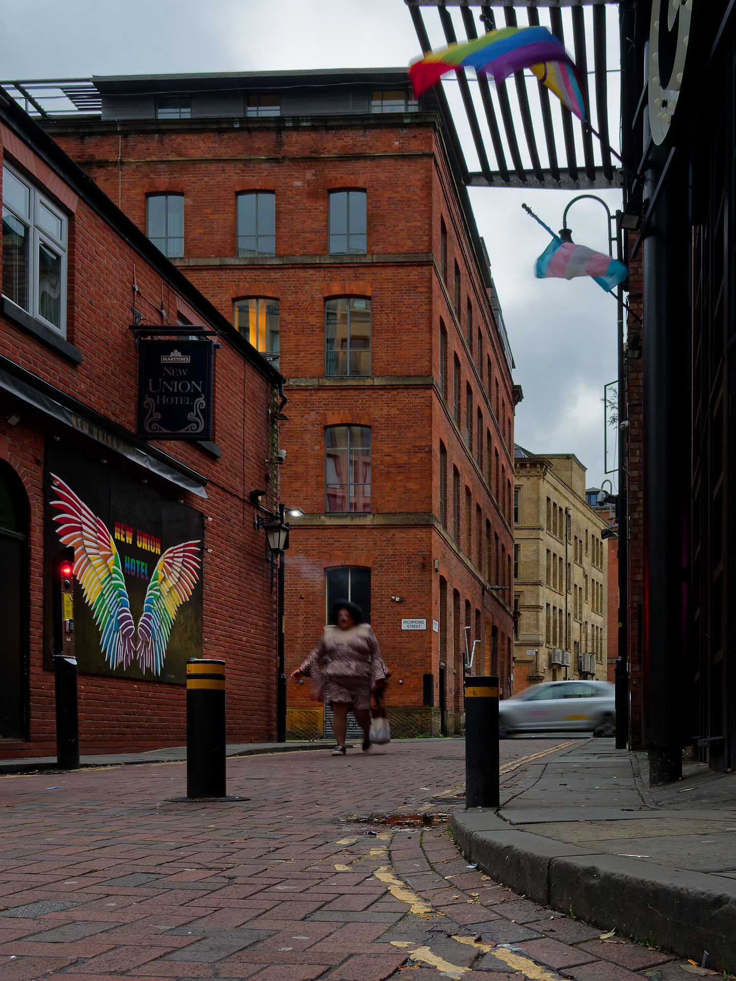 Manchester Manchester Straßenszene: Backsteingebäude, Regenbogenflagge, Person geht auf Pflasterstraße. New Union Hotel mit Flügel-Wandkunst.