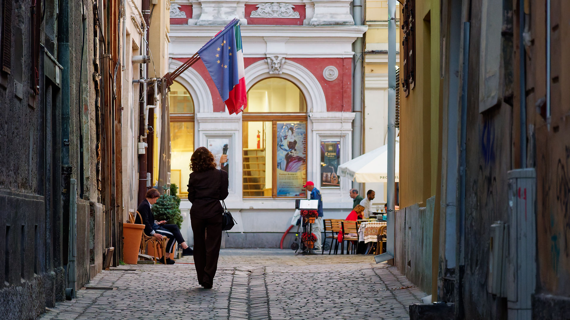 Gasse in Brasov, Rumänien. Frau geht auf Kopfsteinpflaster zu Restaurant mit Europa- und Italienflagge.