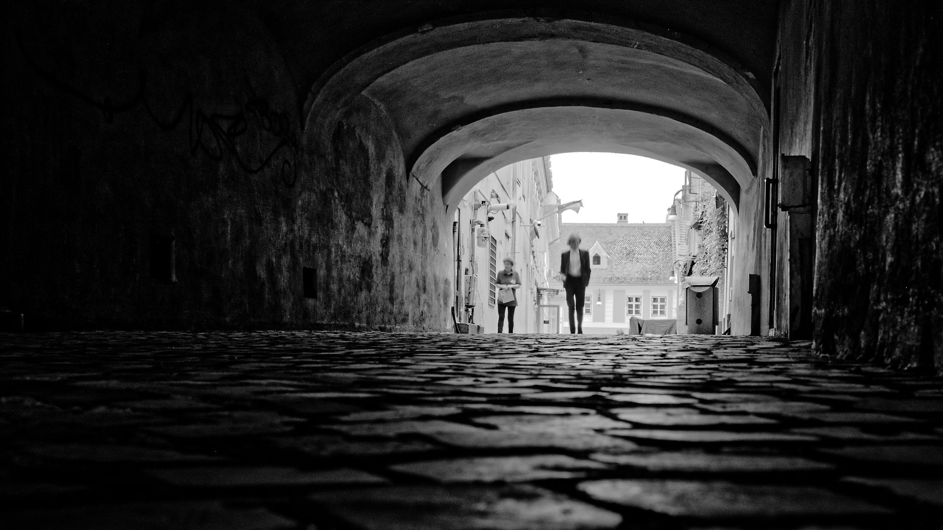 Schwarzweißaufnahme: Gasse in Brasov, Rumänien. Kopfsteinpflaster, Tunnelblick, Personen im Hintergrund. Architektur.
