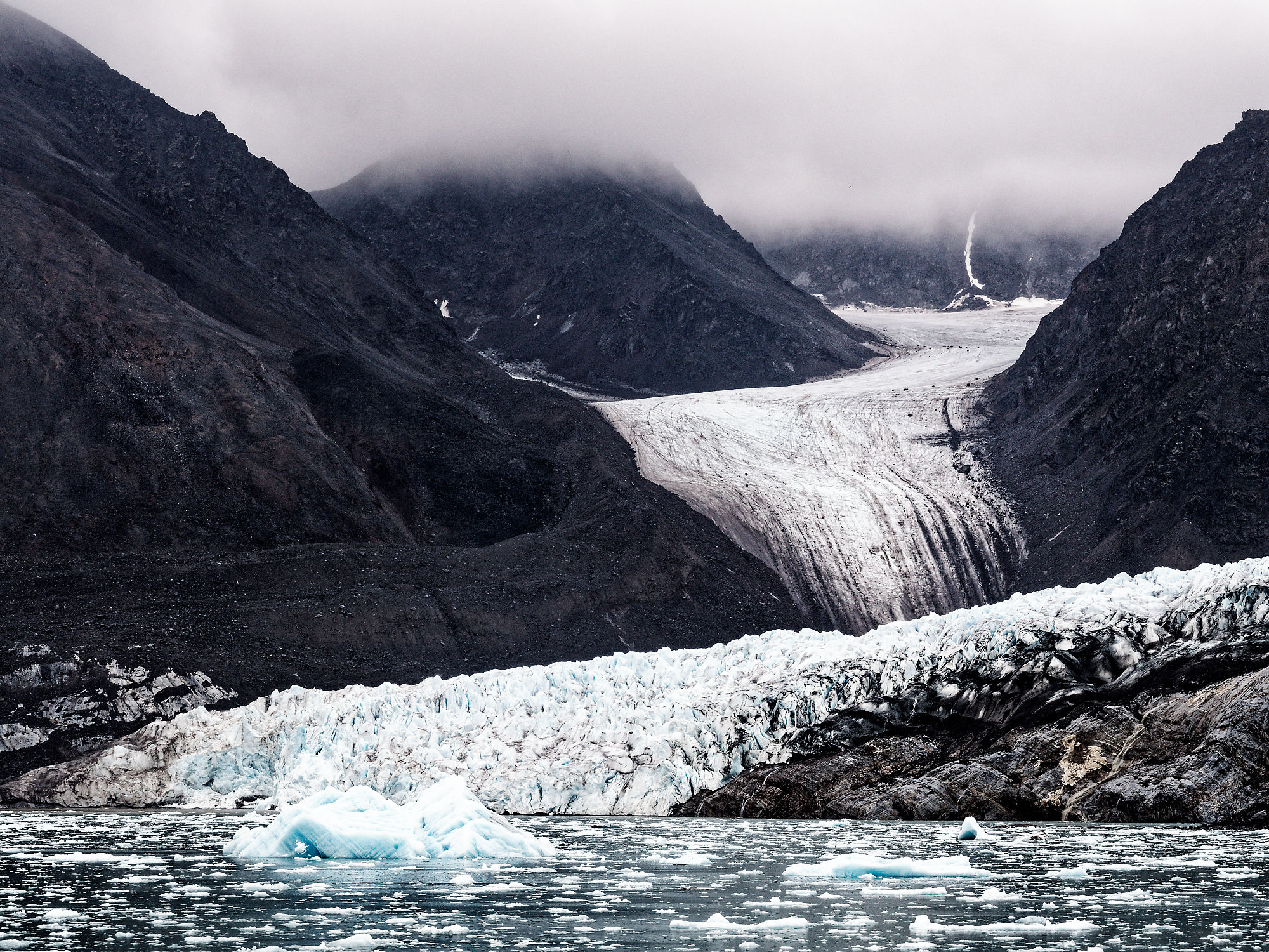 Kongsfjorden Gletscher, Spitzbergen. Eisberge im Wasser vor dunklen Bergen unter bewölktem Himmel.
