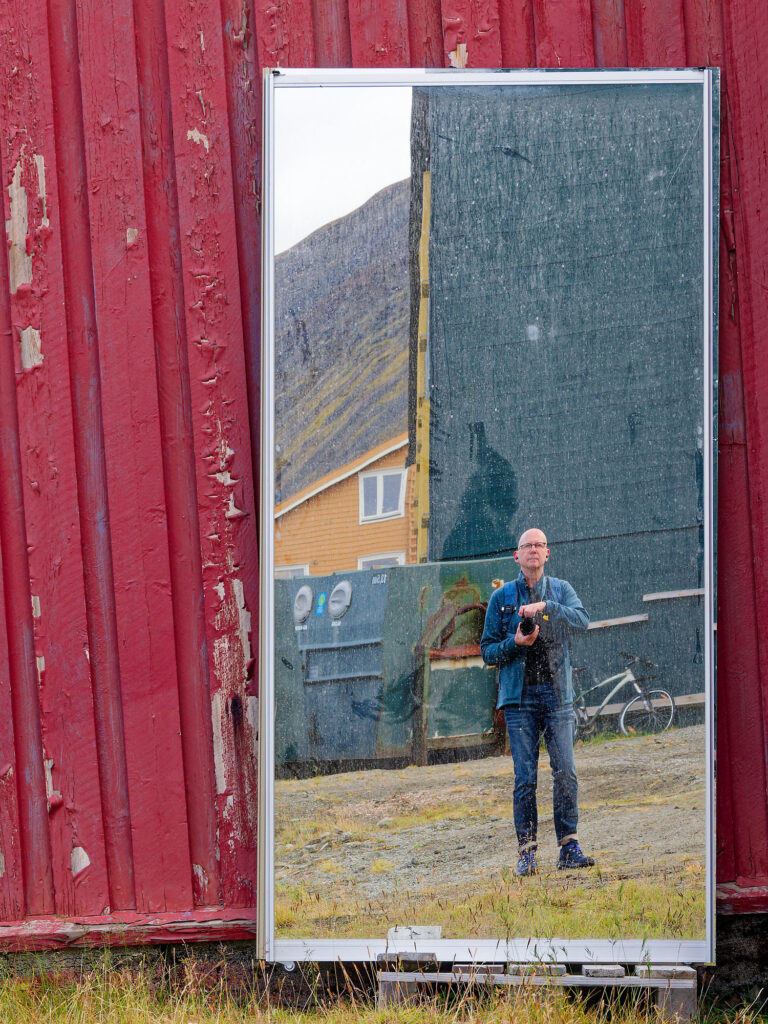 Spiegelbild eines Fotografen in Longyearbyen, Norwegen. Rote Wand, Berg und Haus im Hintergrund. Arktische Fotografie.