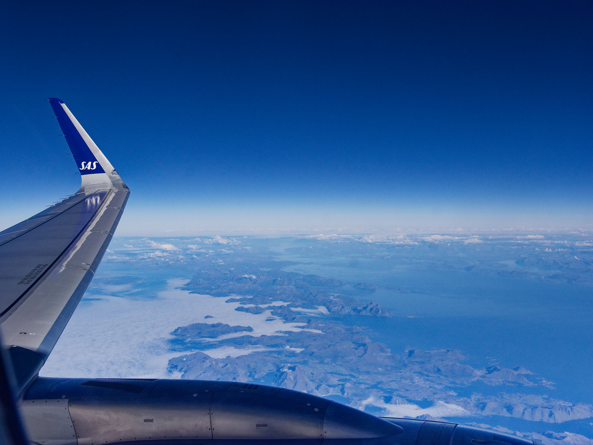 Flugzeugfenster-Ansicht: SAS-Flügel über Küstenlandschaft und blauem Himmel. Flugreise-Impressionen.