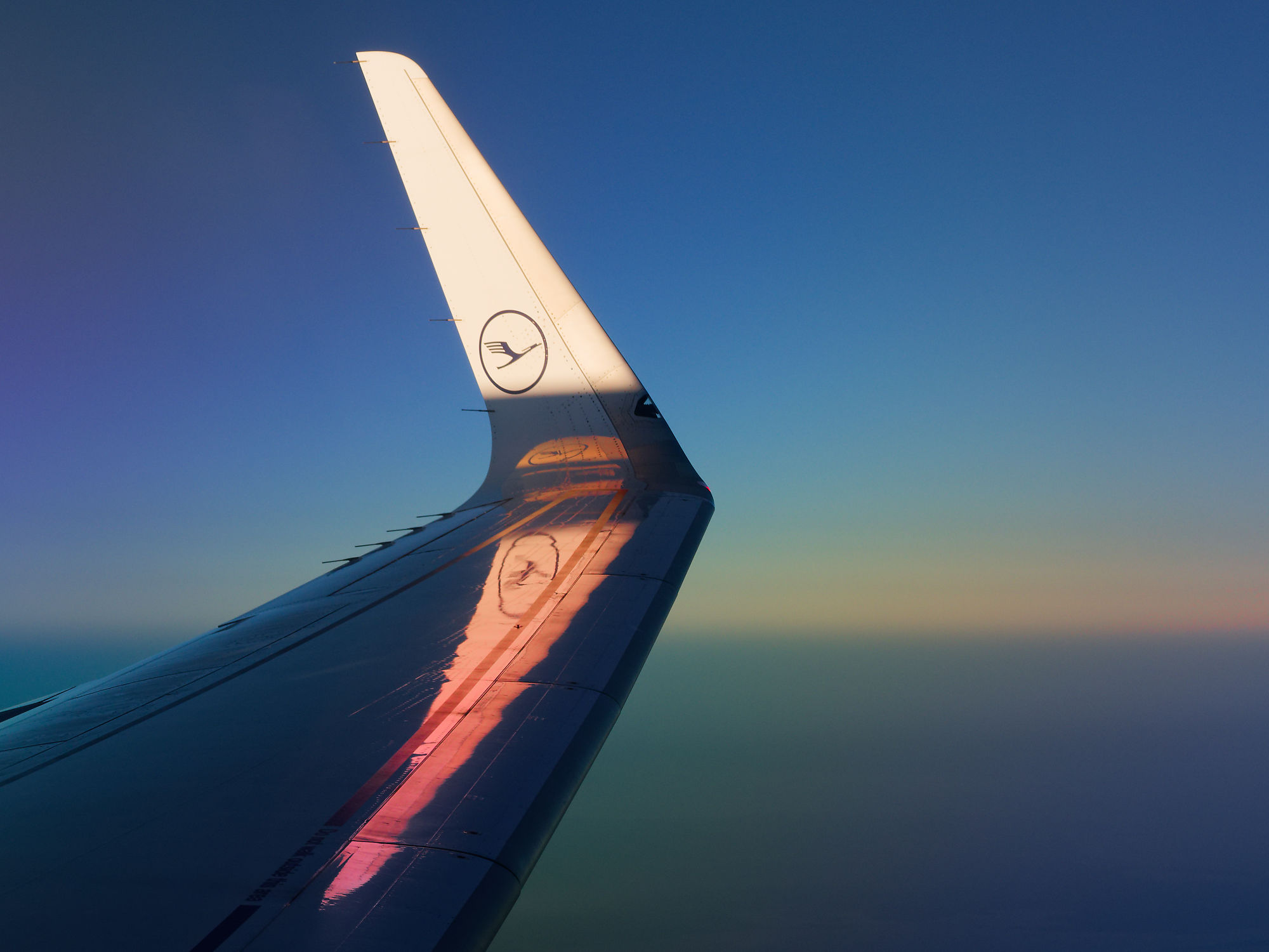 Flugzeugflügel im Flug mit Lufthansa Logo vor blauem Himmel. Logo spiegelt sich auf der Tragfläche.