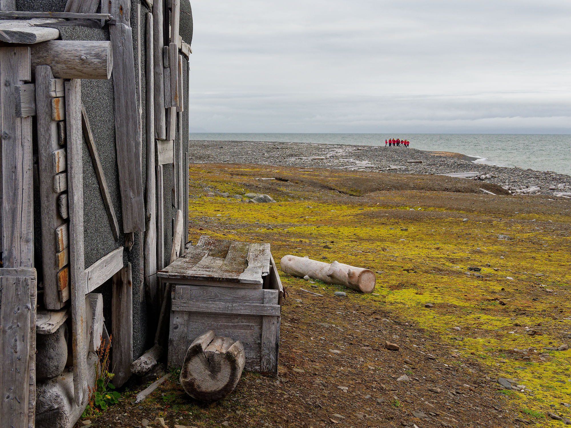 Holzhütte am Woodfjorden, Spitzbergen. Küstenlandschaft mit Moos, Steinen und Personen am Horizont. Arktische Szene.